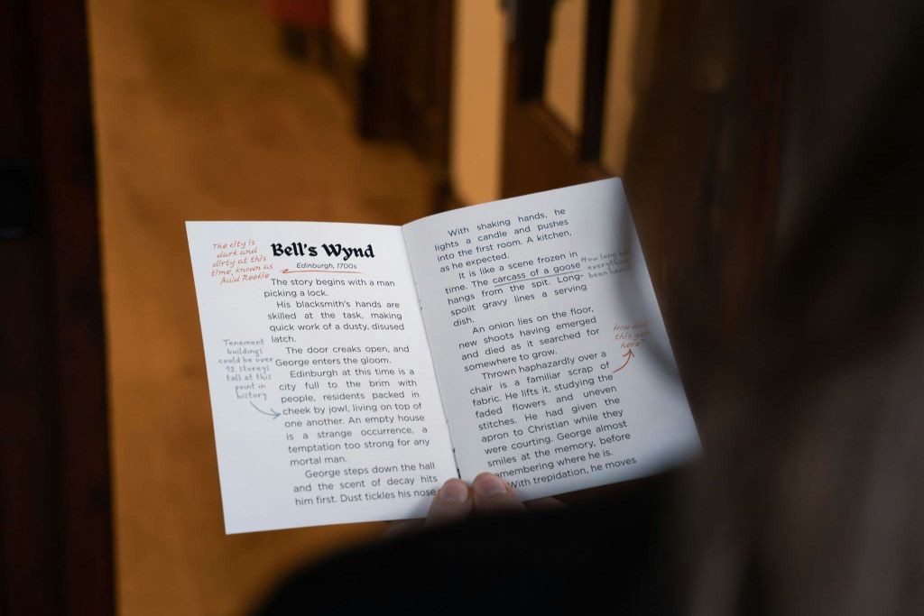 '종의 윈드 (Bell's Wynd)' 라는 이름의 열린 소책자를 들고 있는 사람으로, 인쇄된 텍스트와 손으로 쓴 노트가 빨간색 잉크로 페이지에 새겨져 있습니다.