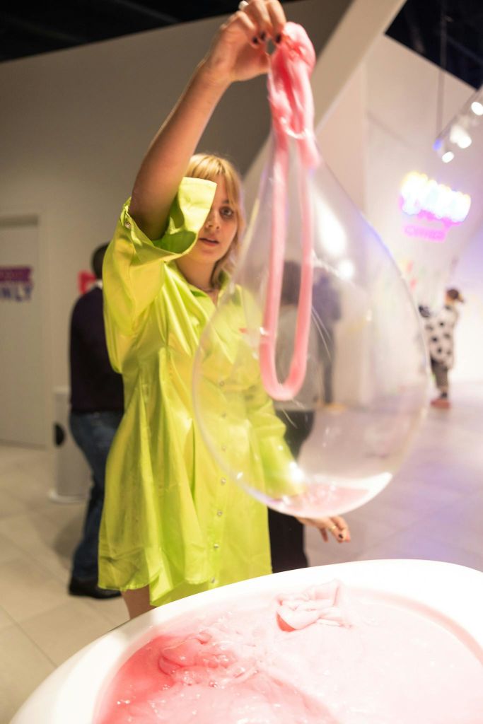 노란색 옷을 입은 사람이 다른 사람들과 네온사인이 있는 밝은 방에서 크고 투명한 풍선과 상호작용합니다.