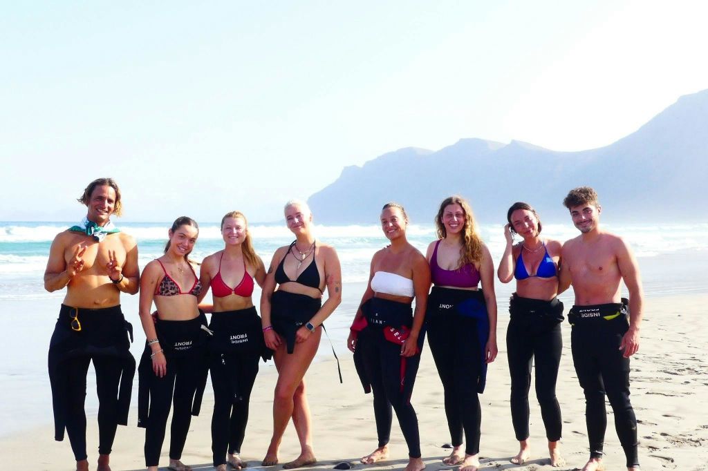 수영복과 잠수복 바지를 입은 6명이 산을 배경으로 해변에 서 있습니다.