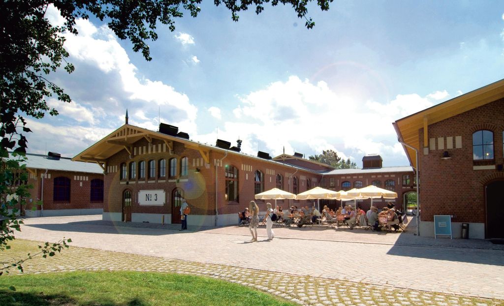 우산 아래 테이블에 앉아 있는 사람들이 있는 햇살이 내리쬐는 마당으로, '3호' 라고 적힌 유서 깊은 벽돌 건물과 인접해 있습니다.