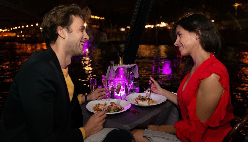 블레이저와 노란색 셔츠를 입은 한 남성이 식사를 하는 동안 미소를 짓고 있으며, 바닷가 도시 풍경과 보라색 조명이 배경을 이루고 있습니다.