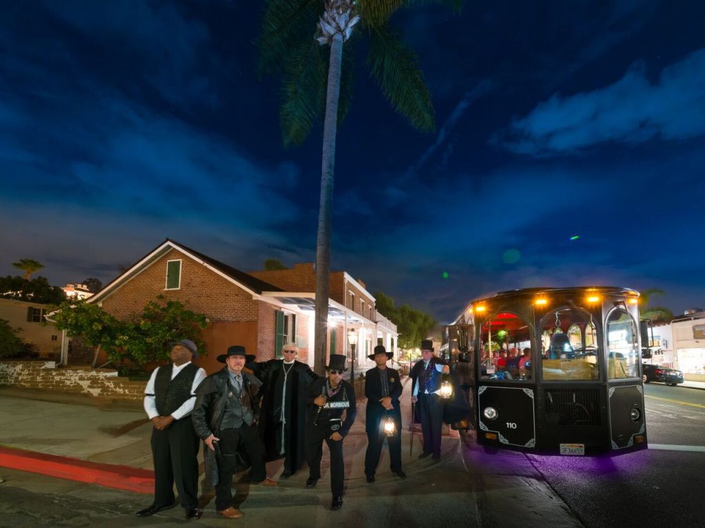 밤에는 벽돌 건물과 야자수 근처의 보도에 마침표를 찍은 옷을 입은 한 무리의 남성이 서 있습니다.