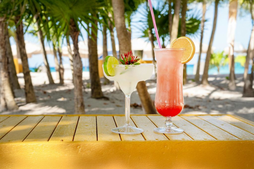 해변과 야자수를 배경으로 노란색 테이블 위에 칵테일 두 잔이 놓여 있습니다. 하나는 라임으로 장식되어 있고, 다른 하나는 오렌지 조각으로 장식되어 있습니다.