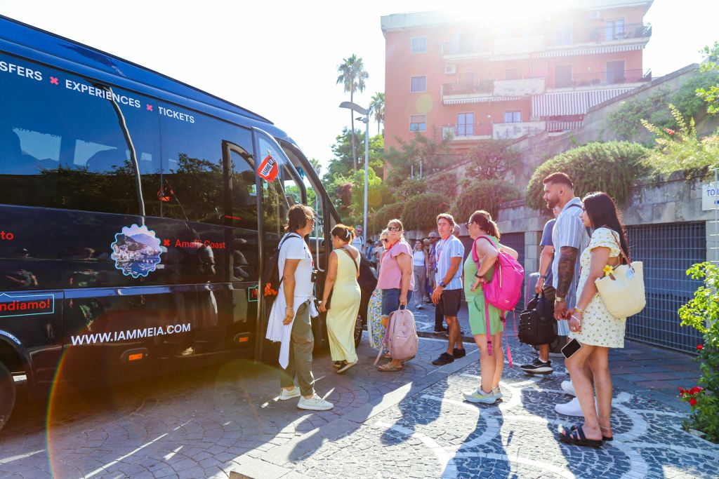 화창한 날, 건물과 녹지를 배경으로 버스에 탑승하는 가방을 든 사람들.