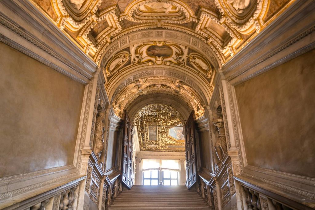 정교하게 조각된 천장, 열린 이중 문, 상단에 빛이 넘쳐흐르는 커다란 창문이 있는 화려한 계단.