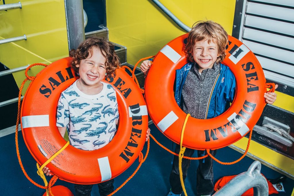 보트 갑판에 서서 "바다의 꿈" 이라고 쓰여진 밝은 주황색 구명 부표를 들고 카메라를 보며 웃고 있는 두 어린이.