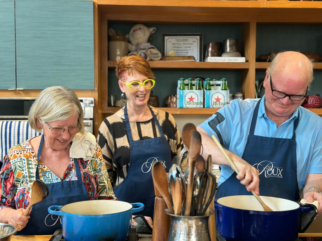 앞치마를 두른 세 사람이 주방에서 함께 요리하고, 냄비, 식기, 물품 선반을 배경으로 요리합니다.
