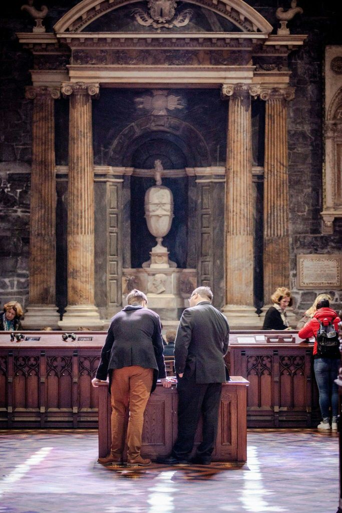 여러 사람이 높은 기둥과 중앙 동상이 있는 역사적인 건물 내부에서 유물을 관찰합니다.