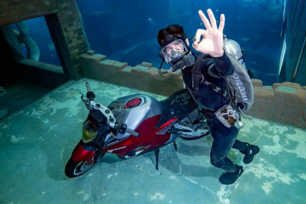 물에 잠긴 구조물로 둘러싸인 'OK' 핸드 사인으로 제스처를 취하는 빨간색 오토바이 옆의 수중 다이버.