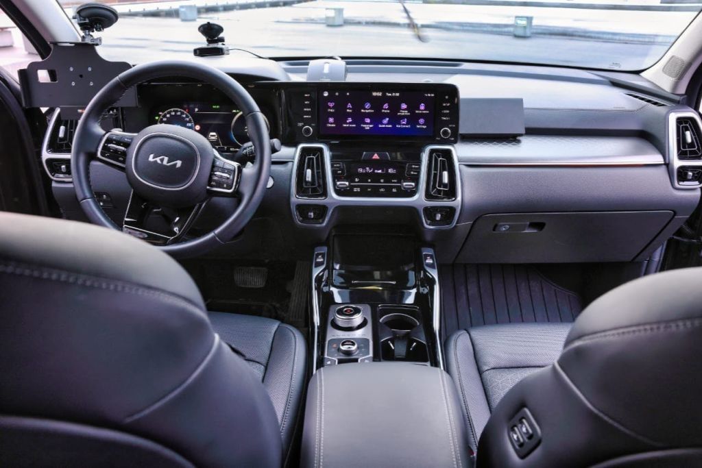 운전석이 보이는 차량 내부, 기아 로고가 있는 스티어링 휠, 디지털 디스플레이가 있는 대시보드, 중앙 콘솔.