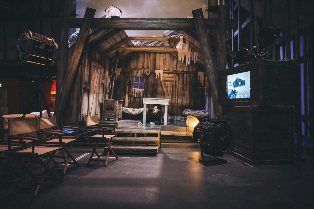 고드름이 있는 오래된 목조 주택을 닮은 소박한 무대 세트. 한 배우가 테이블 아래를 엿보고 있습니다. 영화 장비와 의자가 보입니다.