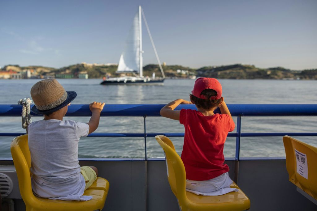 두 명의 어린이가 보트에 앉아 언덕을 배경으로 잔잔한 물가에서 멀리 떨어진 범선을 바라보고 있습니다.