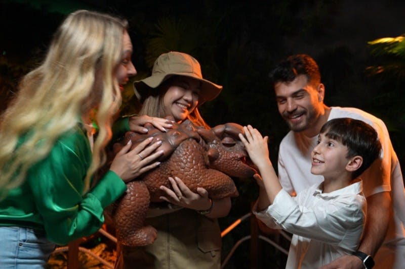 한 무리의 사람들이 미소 짓고 상호작용하고 있으며, 사파리 모자를 쓴 사람이 들고 있는 장난감 공룡을 아이가 만지고 있습니다.