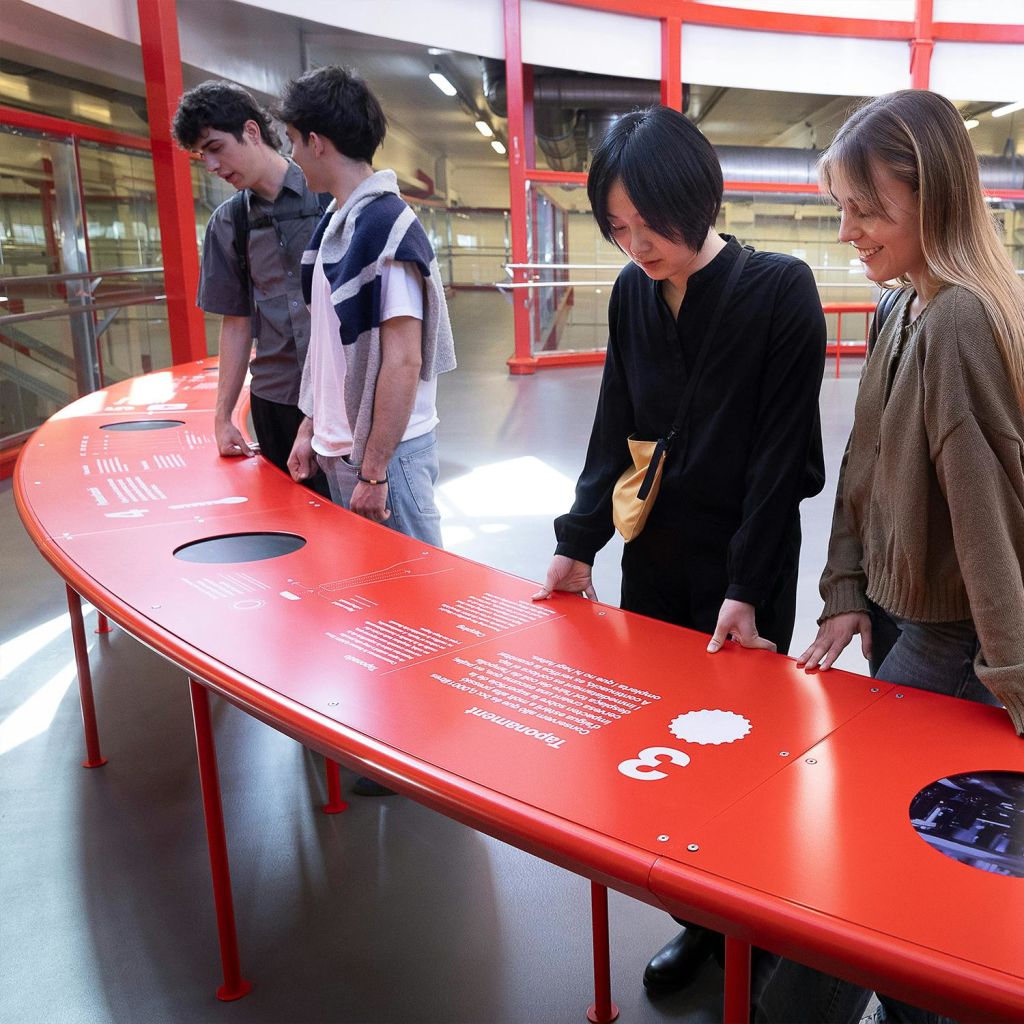 빨간색 구조 요소가 있는 현대적인 건물 내부의 곡선형 빨간색 안내 디스플레이 테이블 주위에 4명이 서 있습니다.