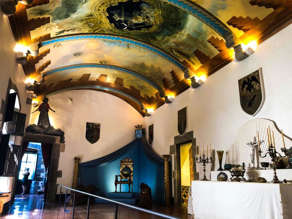 아치형 천장, 화려한 왕좌, 벽 방패, 양초 홀더, 장식된 테이블이 있는 객실입니다.