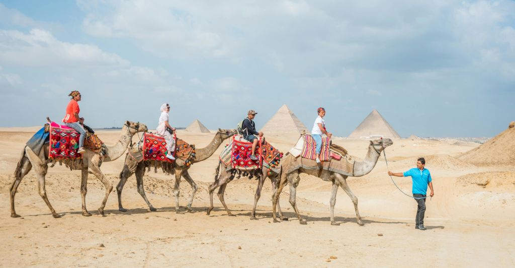 사막에서 화려한 색상의 안장을 쓰고 낙타를 타는 세 사람, 흐린 하늘 아래 피라미드를 배경으로 한 사람들.