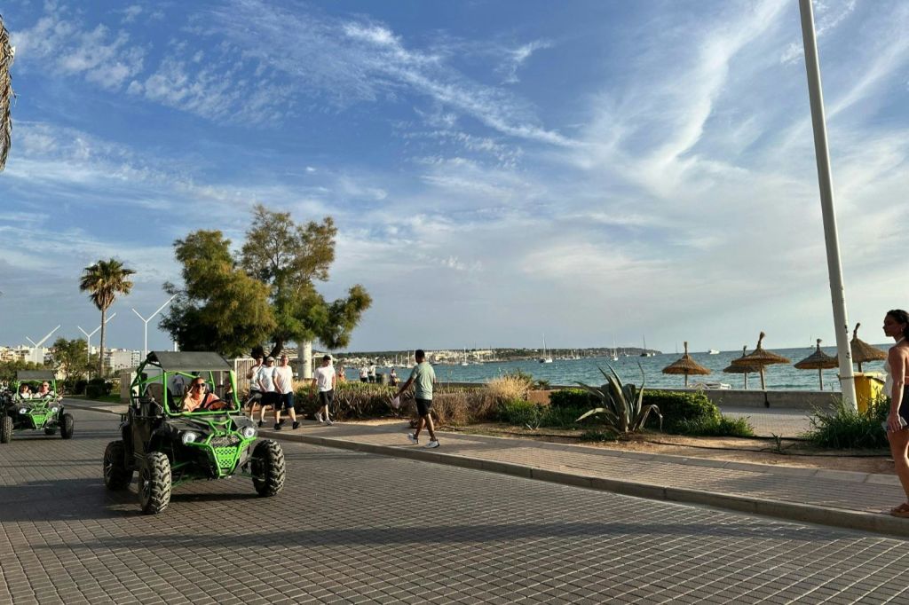 통로, 녹색 모래 언덕 버기, 나무, 우산, 부분적으로 흐린 하늘 아래 바다가 있는 해변 근처를 걷는 사람들.