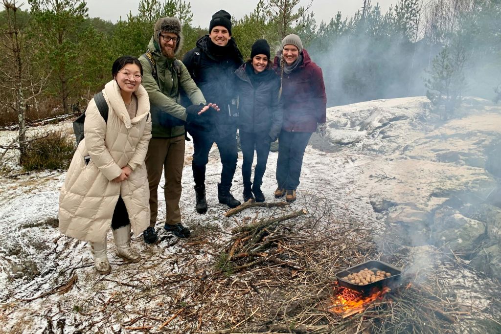 겨울 옷을 입은 다섯 명이 눈으로 뒤덮인 숲 속 작은 모닥불 주위에서 미소를 짓고 서 있습니다.