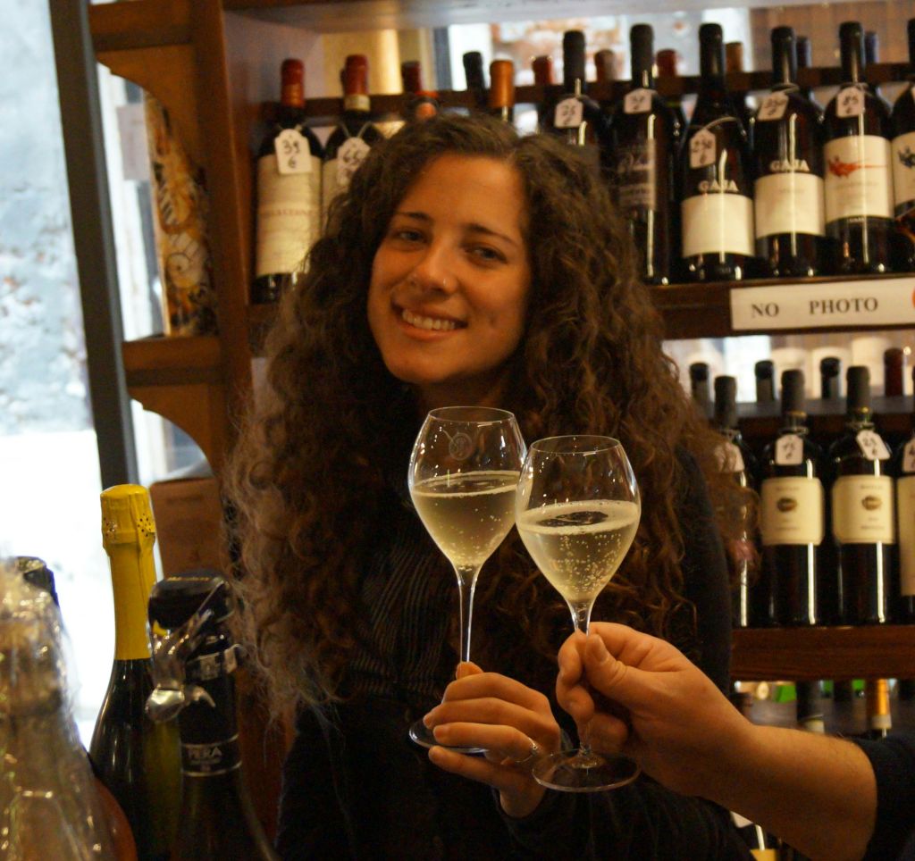 곱슬머리를 한 여성이 주류 매장에서 화이트 와인 한 잔을 들고 미소를 짓고 있습니다. 또 다른 사람이 그녀와 함께 술잔을 움찔거렸다.