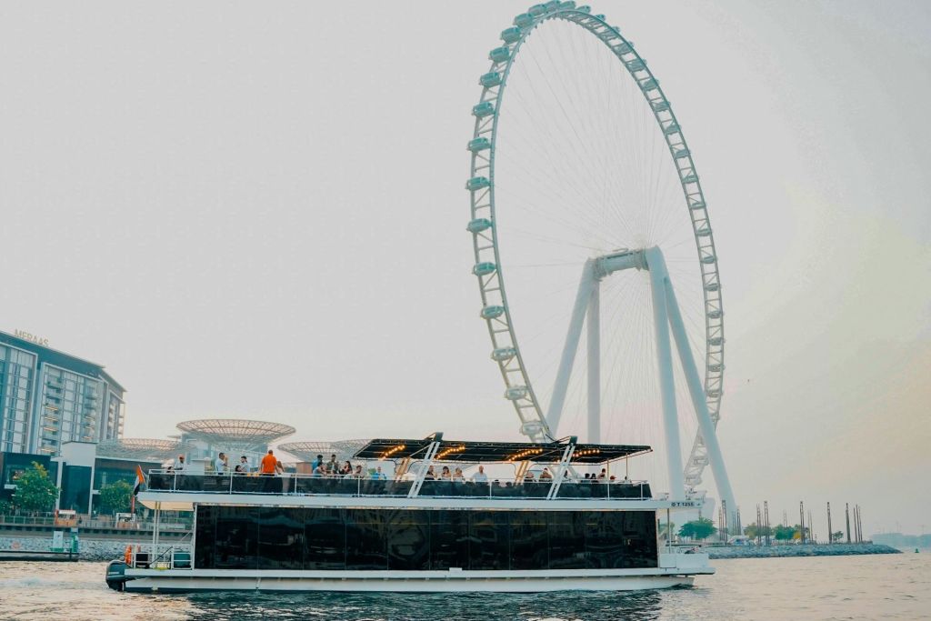 상부 갑판에 많은 사람들이 타고 있는 보트가 커다란 전망대 근처의 물 위에 떠 있습니다.