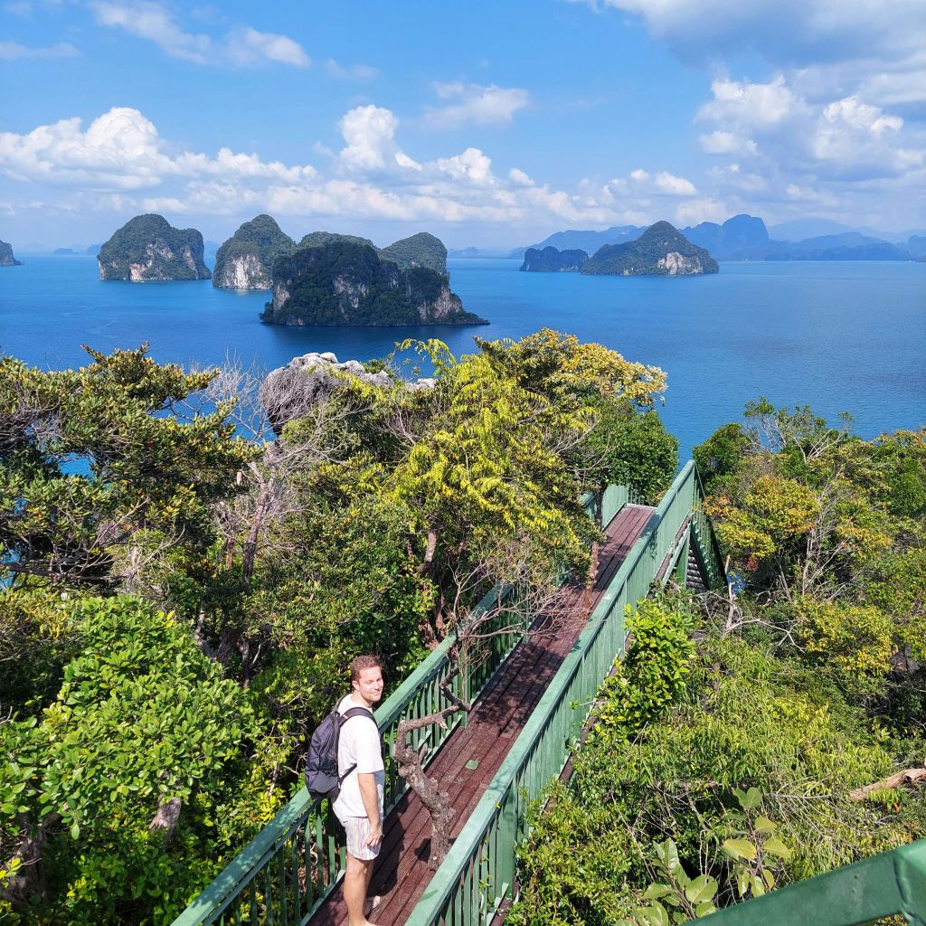 조그만 바위섬이 펼쳐진 푸른 바다가 내려다보이는 빽빽한 녹음 속에 배낭을 든 남성이 나무 산책로에 서 있습니다.