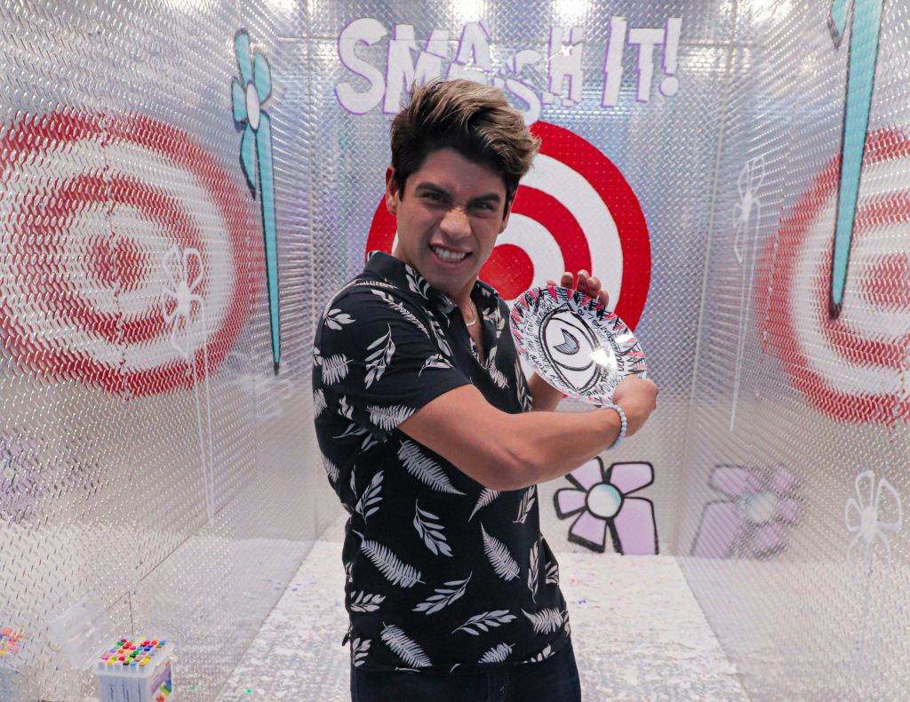 검은색 무늬의 셔츠를 입은 사람이 깨진 접시를 들고 위에 적힌 "Smash IT!" 가 있는 표적 앞에서 포즈를 취하고 있습니다.