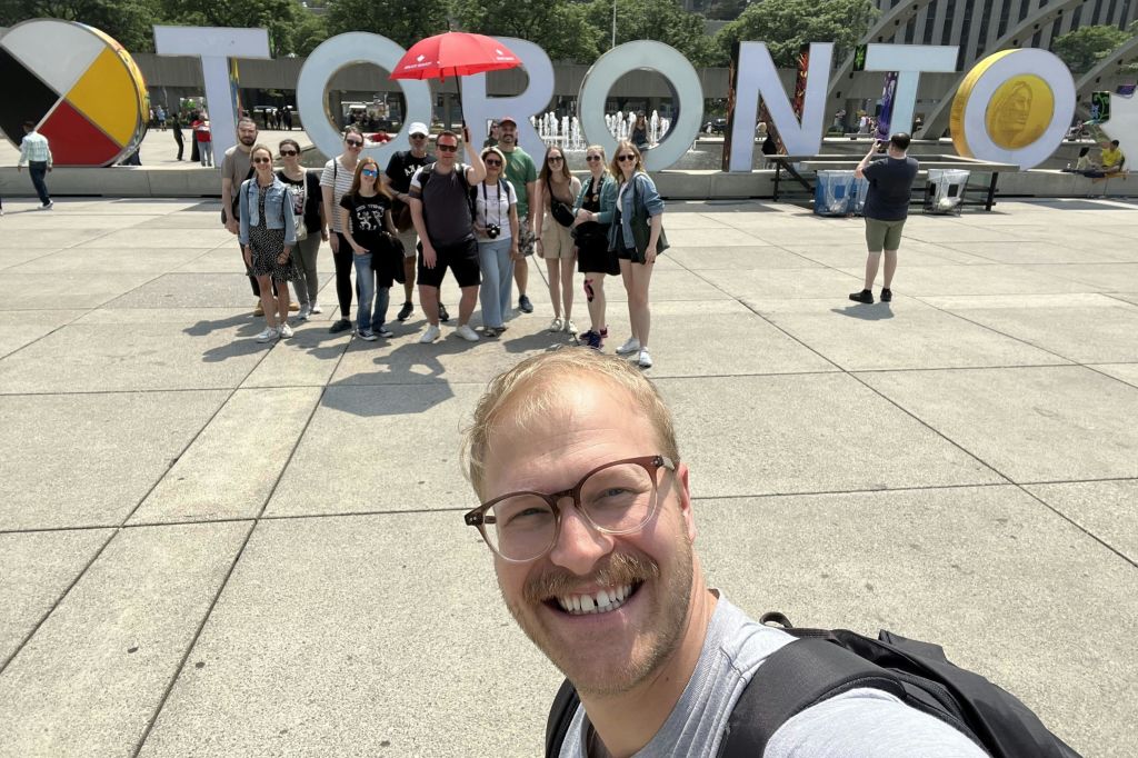한 무리의 사람들이 광장에서 커다란 "토론토" 편지 앞에서 포즈를 취하고 있으며, 맨 앞자리에 있는 한 사람이 셀카를 찍고 있습니다.