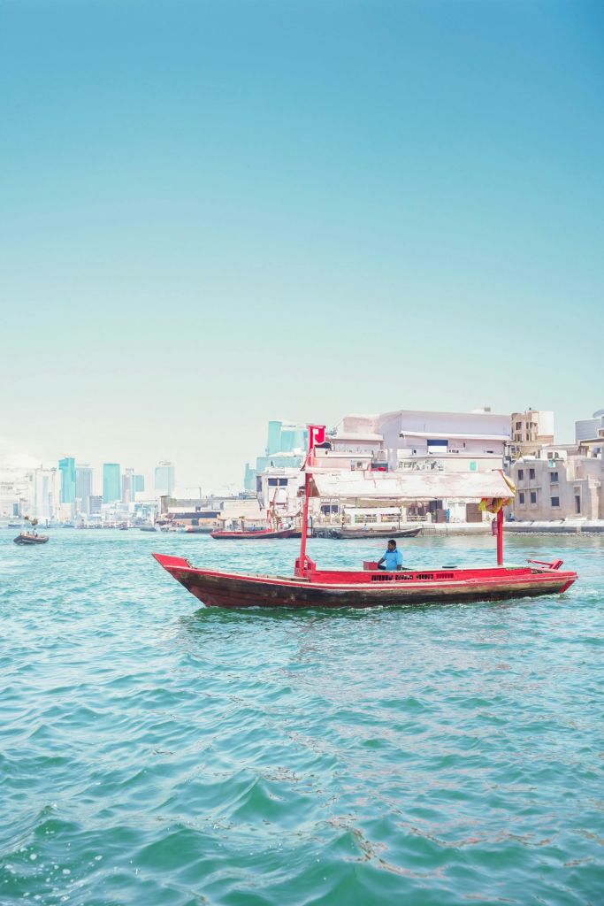 도시의 스카이라인과 수변 건물을 배경으로 화사하고 화창한 날에 전통적인 붉은 보트를 조종하는 사람.