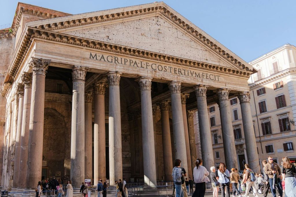 로마의 판테온 앞에 서 있는 사람들은 커다란 기둥과 페디먼트에 새겨진 문구가 있습니다.