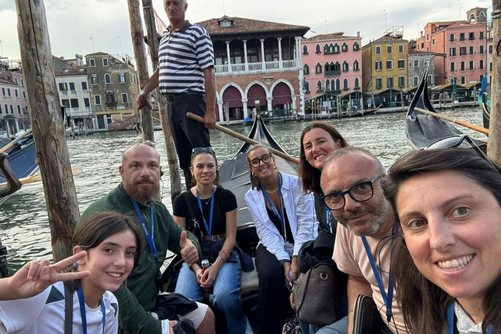 곤돌라를 탄 7명의 일행이 곤돌라 뒤에 서 있습니다. 운하를 배경으로 베네치아 양식의 건물들이 늘어서 있습니다.
