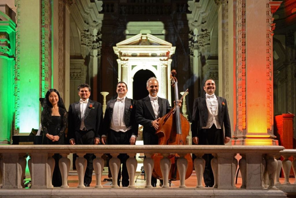 녹색 조명과 흰색 아치를 배경으로 화려한 건축 장식이 돋보이는 무대에 정장 차림의 뮤지션 5명이 서 있습니다.