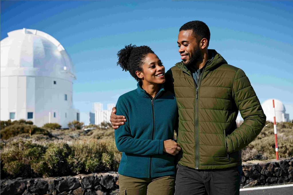 화창한 날씨에 재킷을 입은 미소 짓는 커플이 화분과 건물을 배경으로 야외를 거닐고 있습니다.