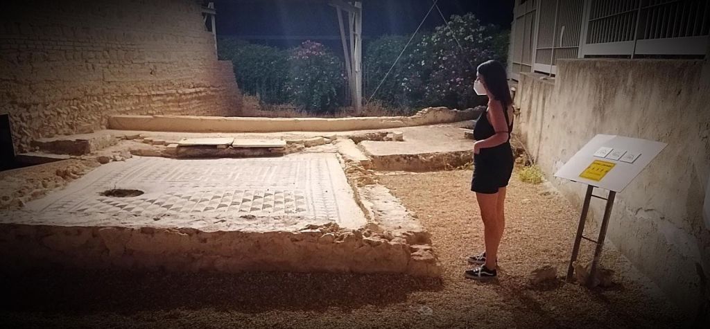 검은색 드레스와 얼굴 마스크를 쓴 여성이 타일 바닥이 있는 고대 유적 옆에 서 있으며, 밤에는 식물과 조명으로 둘러싸여 있습니다.