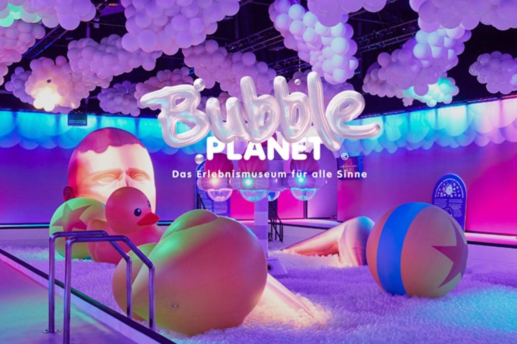 거대한 고무 오리, 다채로운 풍선, 네온 조명 아래 매달린 풍선이 있는 생동감 넘치는 전시회입니다. 텍스트는 "Bubble Planet" 입니다.