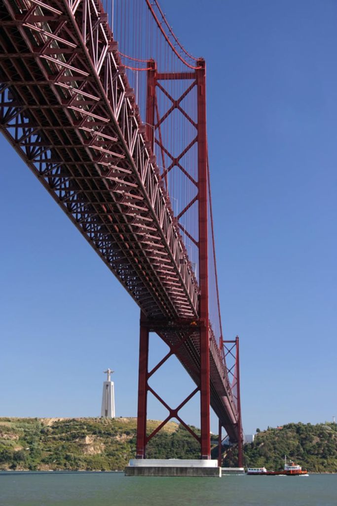 맑고 푸른 하늘을 배경으로 수평선에 그리스도의 동상이 있는 커다란 붉은 현수교 아래에서 바라보는 풍경.