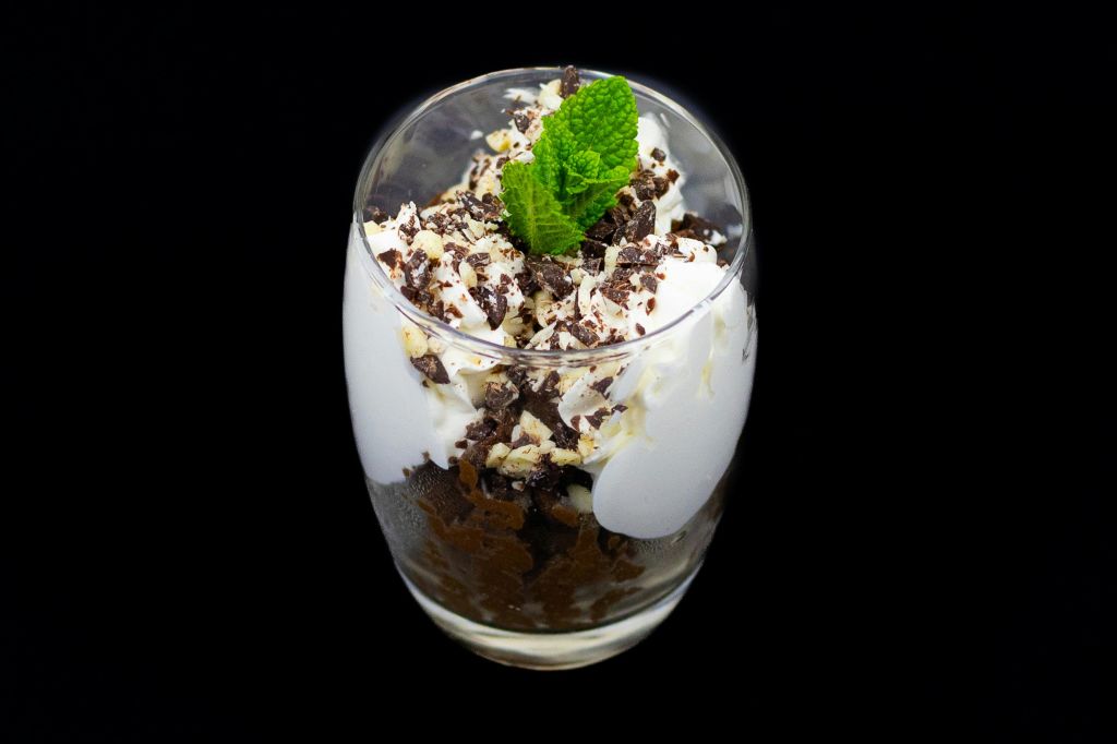 검은색 배경에 휘핑 크림, 초콜릿 쉐이빙, 민트 장식이 들어간 잔에 담긴 디저트.