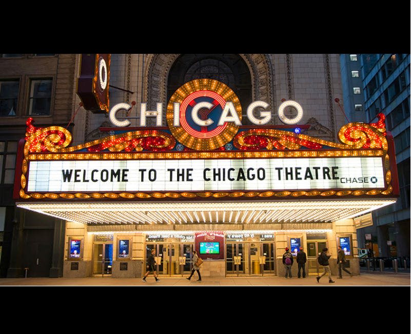 시카고 극장의 조명이 켜진 후미에는 "시카고 극장에 오신 것을 환영합니다" 라는 메시지가 표시되어 있으며, 아래를 걷는 사람들이 있습니다.