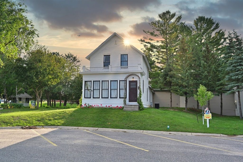 푸른 나무와 잔디밭으로 둘러싸인 현관이 있는 2층 화이트 하우스로 전경에 주차장이 있습니다.