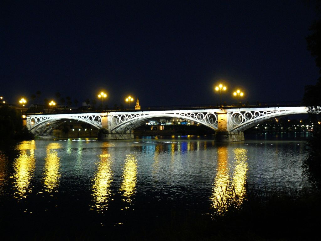밤에는 아치형 다리에 불이 들어오고, 아래 물에 빛이 반사되며, 도시 풍경이 배경으로 보입니다.
