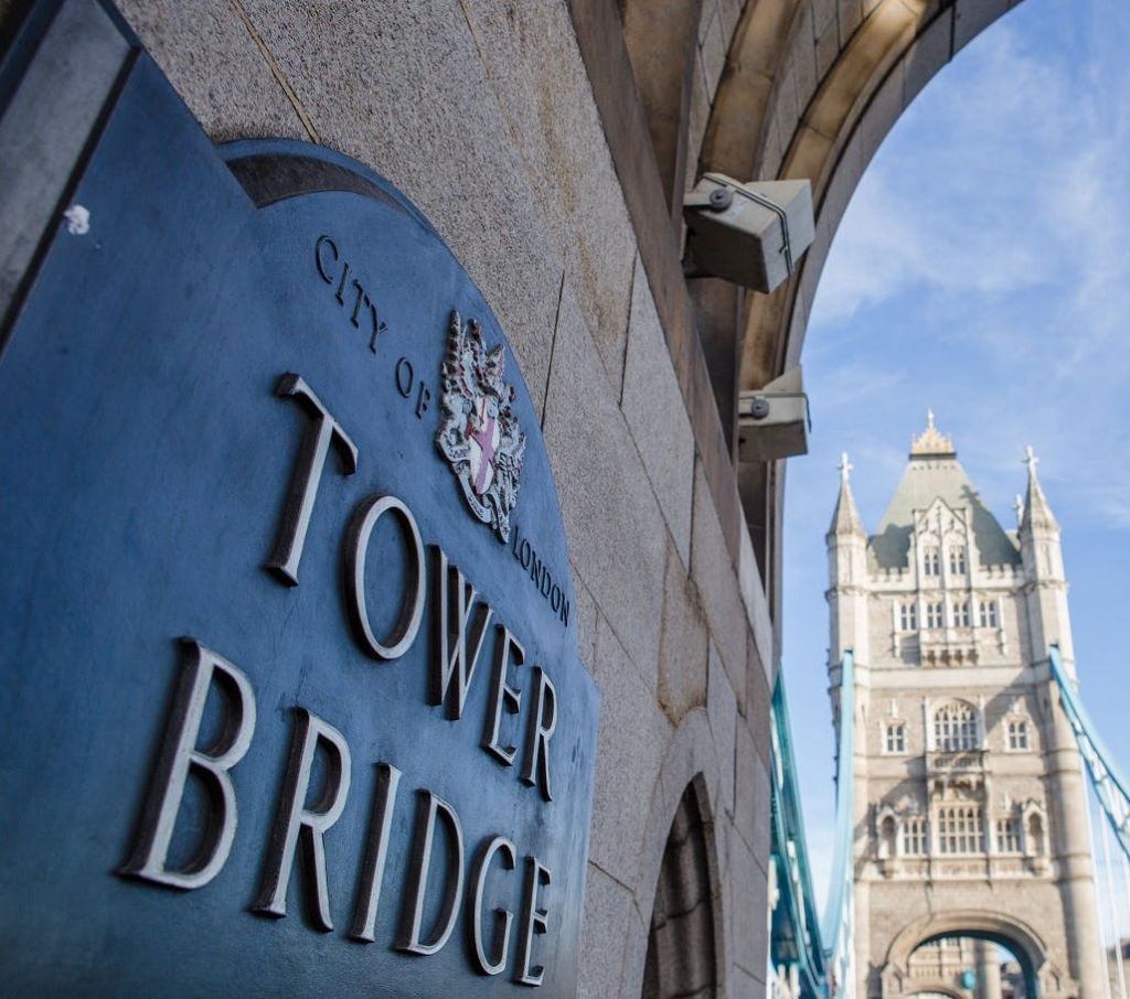 맑은 날 타워 브리지를 배경으로 아치형 통로 아래 '시티 오브 런던, 타워 브리지' 라고 적힌 간판.