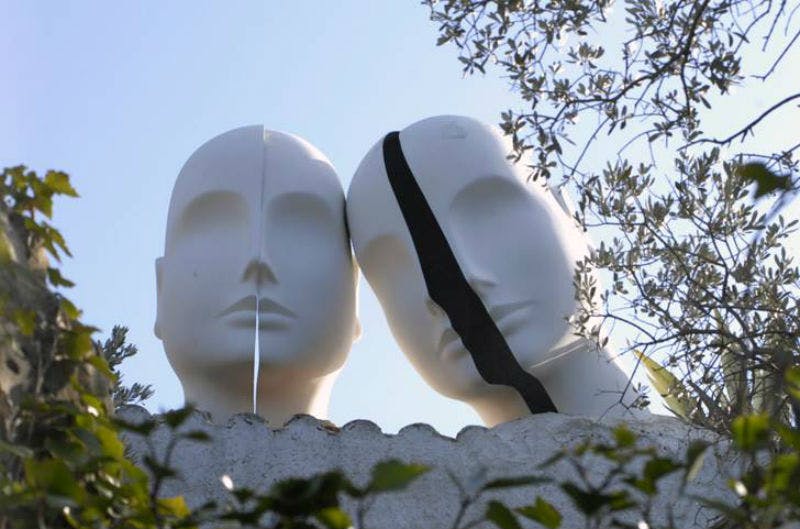 얼굴이 갈라진 두 개의 마네킹 머리, 하나는 흰색 줄무늬가 있고 다른 하나는 검은색 줄무늬가 있으며 파란 하늘과 단풍을 배경으로 합니다.