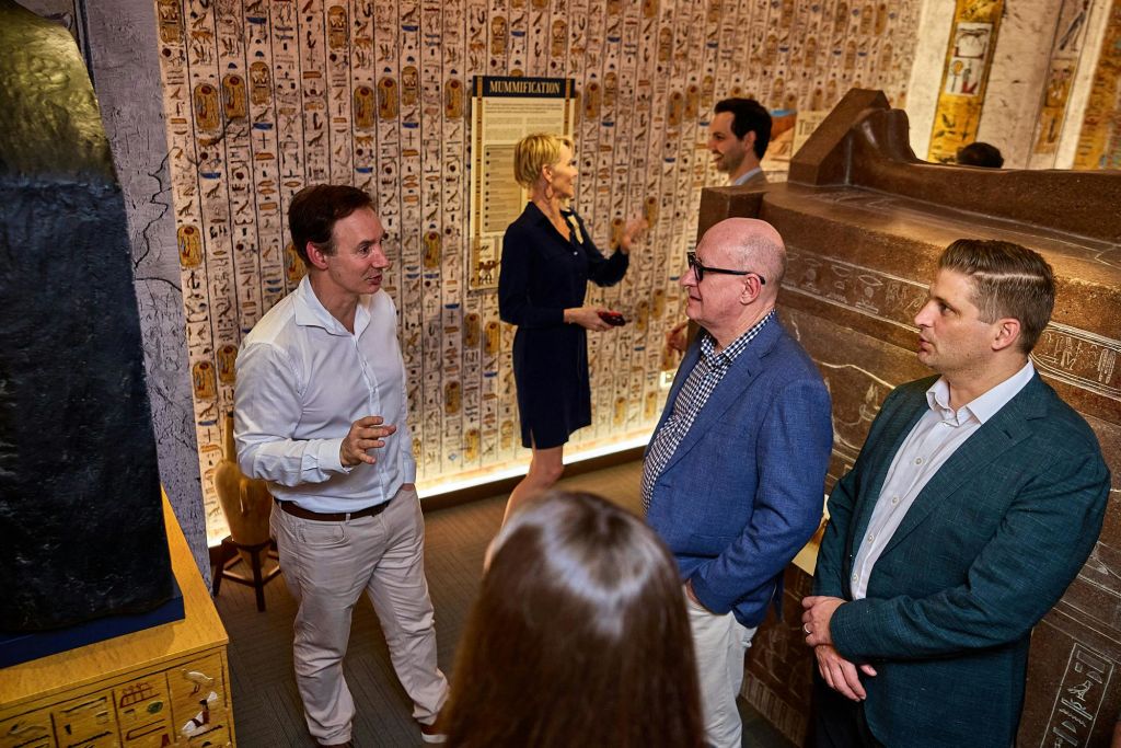 고대 이집트 장식과 미라를 배경으로 방에서 대화하는 정장 차림의 사람들.