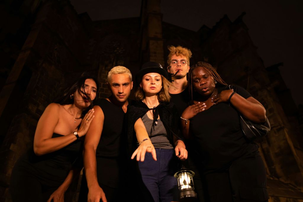등불이 켜진 어두운 옷을 입은 5명의 사람들이 밤에 고딕 양식의 건물을 뒤로 한 채 바짝 붙어 서 있습니다.