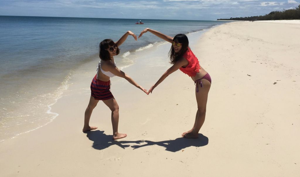 햇살이 내리쬐는 해변에서 두 명의 여성이 팔로 하트 모양을 만들어 멀리 떨어진 패들보더와 함께 해안가 근처에서 미소를 짓고 포즈를 취하고 있습니다.