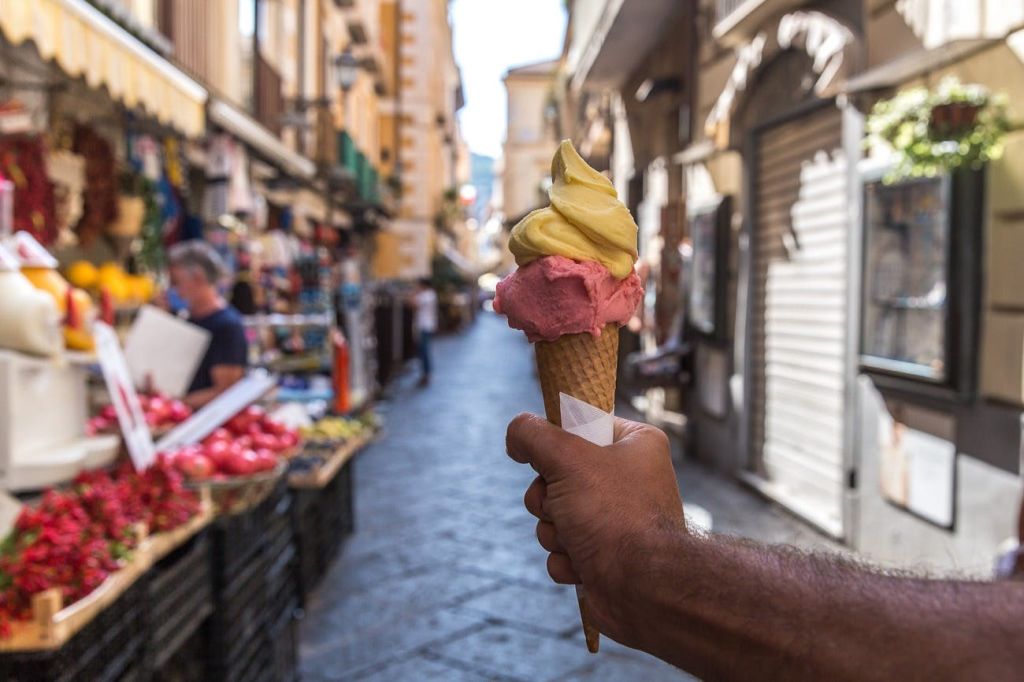 상점과 사람들을 배경으로 좁고 번화한 거리에서 노란색과 분홍색 아이스크림이 담긴 콘을 들고 있는 손.
