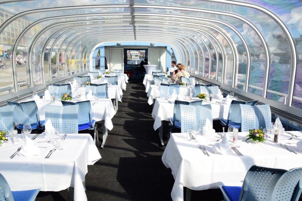 유리 지붕이 있는 레스토랑, 흰색 침구가 놓인 깔끔한 테이블, 파란색 의자, 두 사람이 식사를 하고 있습니다.