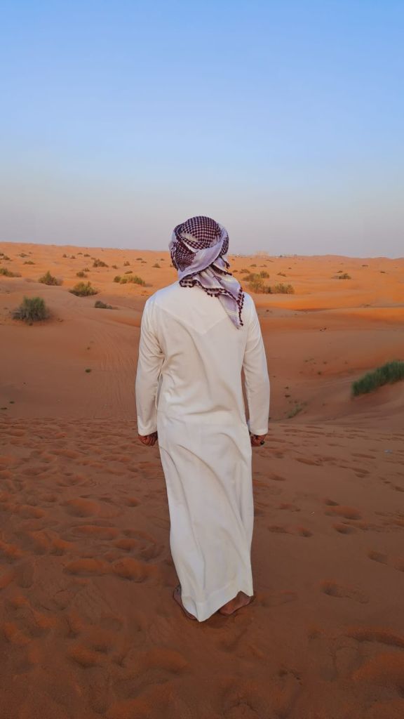 맑은 하늘 아래 희박한 초목이 있는 사막 풍경을 걷는 하얀 가운과 머리 스카프를 입은 사람.