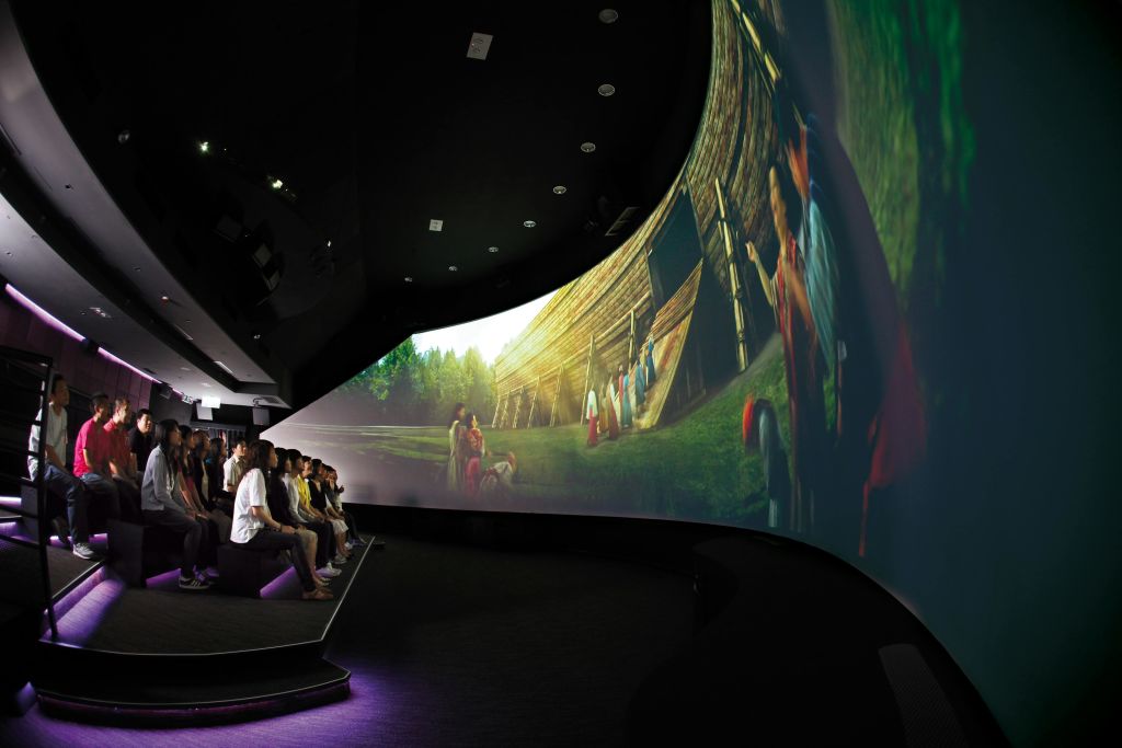 어두운 극장에 앉아 다양한 캐릭터가 등장하는 역사적 재연 장면을 보여주는 커다랗고 곡선형의 스크린을 보는 사람들.