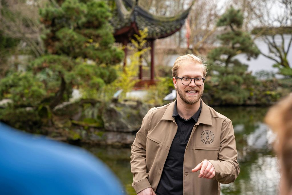 연못과 전통 정자를 배경으로 정원에 서 있는 황갈색 재킷과 안경을 입은 남성.