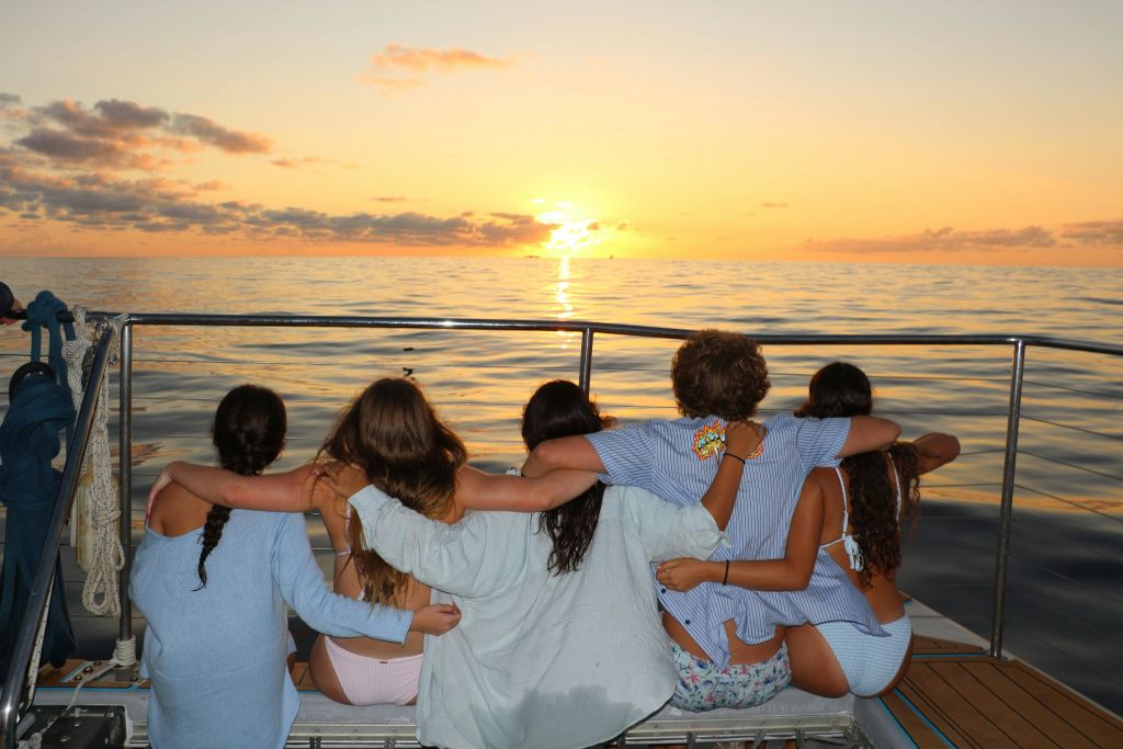 다섯 명이 보트 갑판에 앉아 서로 팔짱을 끼고 잔잔한 바닷물 위로 지는 석양을 바라보고 있습니다.